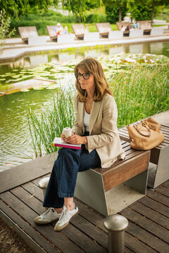 A confident blond haired woman in glasses sitting on a bench in a city park by a pond and holding a coffee cup and a book
