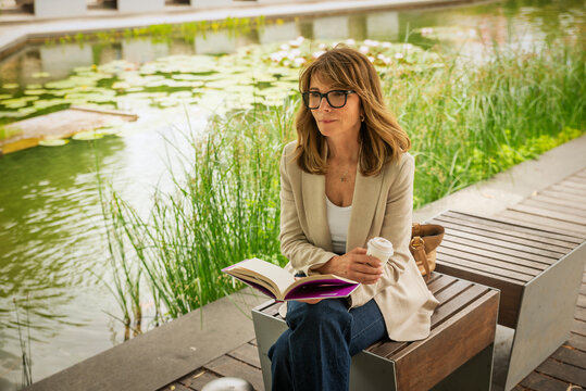Confident blond haired woman reading a book with coffee in a modern city park