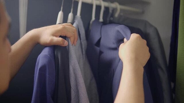 Close up of man hand choosing elegant blue suit jacket from hanging rack in closet