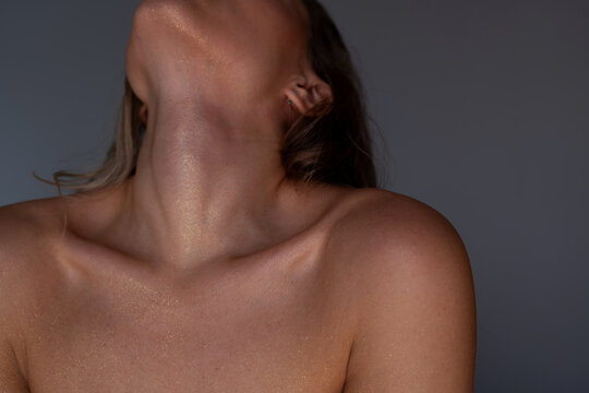 Close-up of young woman neck and collarbones with golden shimmer on glowing skin, sensual beauty concept with soft studio light on a grey background