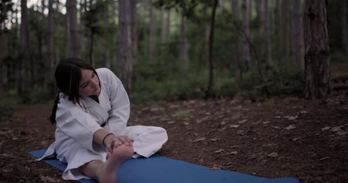 A Young Girl in a White Karate Gi with a Red Belt Performs Stretching Exercises on a Blue Yoga Mat Amidst the Serene Beauty of a Pine Forest, Focusing on Flexibility and Discipline During Her Martial