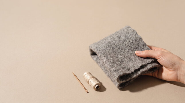 Minimalist craft flat lay of hand holding textured wool felt near spool and needle in warm beige and grey shades representing sustainable textile handicraft with copy space