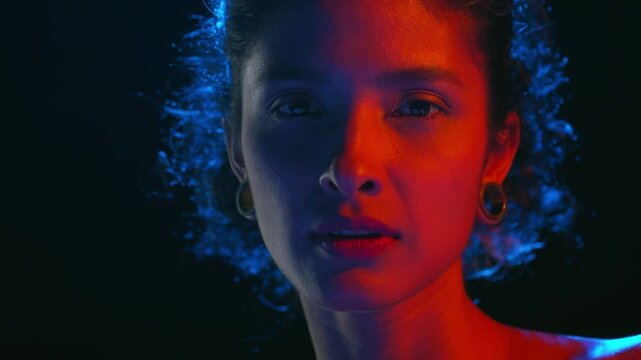 Detailed shot of cheerful latin american model with curly hair illuminated with blue and red turn her body stroking her body skin from back to clavicles against black bg | Fragrance ad concept