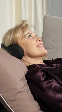 Smiling elderly woman relaxing on a comfortable sofa at home, listening to her favorite song on wireless headphones with her eyes closed, enjoying a peaceful and serene moment of happiness