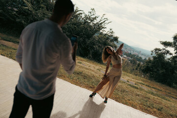 Obraz premium Photographer capturing a female model dancing and posing during an outdoor fashion photo shoot on a sunlit hillside with city view