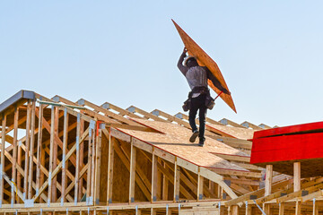 Construction worker carrying an OSB roof sheathing panel on the roof of a two story single family home under construction, representing residential framing, material handling, and housing development © Eduardo Barraza