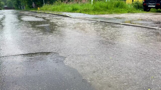 Rainwater collects on the asphalt road, forming puddles with ripples and reflections, surrounded by grass and roadside elements in the background