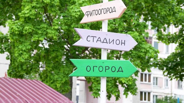 A wooden post features several colorful arrow pointer signs with Russian inscriptions indicating various locations against a background of lush green summer trees.