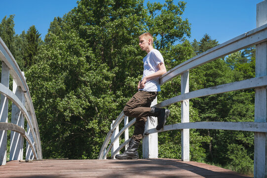 Teenager standing on a white wooden bridge surrounded by green trees, wearing a white T shirt, brown cargo pants and black boots, leaning on the railing under a clear sky