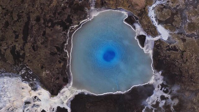 Aerial view of a circular geyser pool in Iceland, deep blue throat, white mineral rim, dark volcanic ground, steaming vents, ripples, and runoff channels visible