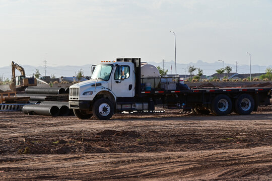 Service flatbed truck at a residential development site with underground utility materials, while an excavator and worker perform dust control during housing infrastructure construction