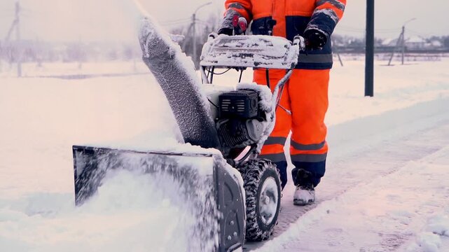 Worker clears snow with powerful snow blower. Orange suit stands out against white winter landscape. Snow flies as machine pushes icy ground. Cold air, steady hands, focused effort