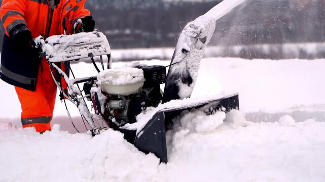 Worker clears snow with heavy machinery. Snow flies as machine cuts path through thick snow. Person wears bright orange jacket for visibility. Winter scene shows routine snow removal task