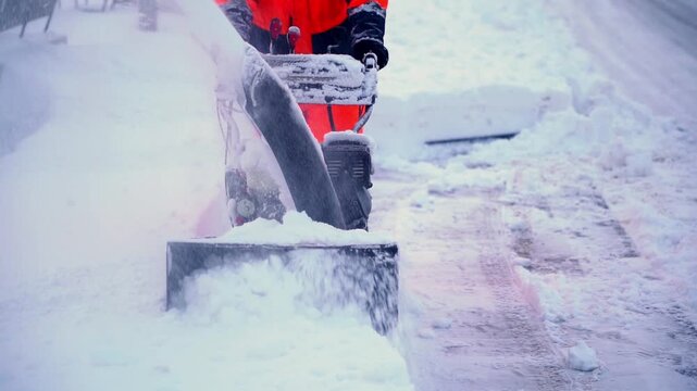 Worker clears snow with heavy-duty snowblower. Snow flurries swirl around machine's blade. Cold air bites as machinery grinds through ice. Worker moves steadily through deep winter debris