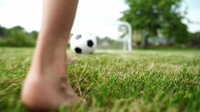 Child's bare feet approach soccer ball on grass. Goalpost stands blurred in background. Scene captures playful outdoor sports moment. Footage evokes carefree, energetic, joyful motion