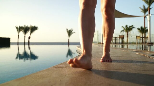Sunlight glows warmly over bare feet stepping near infinity pool. Reflections ripple gently, mirroring palms and sky. Silent steps echo along poolside ledge. Relaxed pace suggests serene escape
