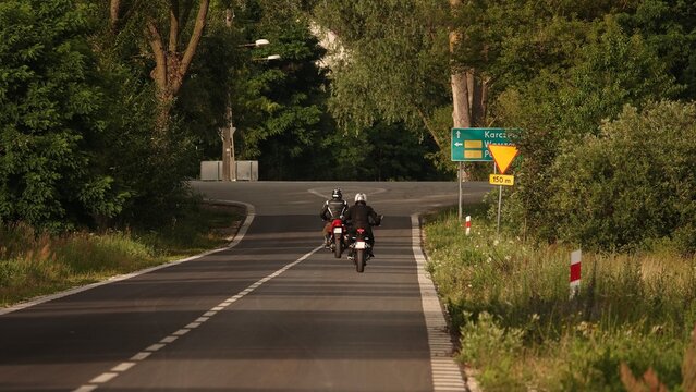 Zdjęcie przedstawia dw&oacute;ch motocyklist&oacute;w jadących asfaltową drogą w stronę skrzyżowania. Trasę otaczają gęste zielone drzewa i krzewy w letnim świetle.