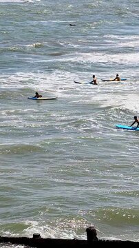 Vertical panning shot of a distant surf school session on Cromer Beach, North Norfolk coast. The camera moves across surfers riding the waves, capturing the seaside setting, blue sea.