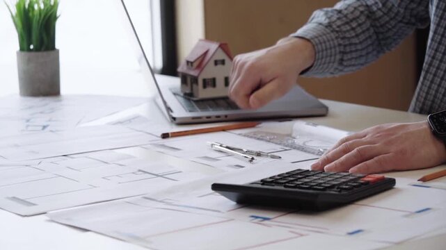 Hand draws blueprint near calculator and mini house model. Focused person plans home project with precision. Workspace holds laptop, pencils, and design sheets. Ideal for real estate, construction