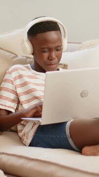 Vertical video: Typing teen male in headphones and striped tee, tapping laptop and studying on sofa