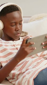 Vertical video: Opening eyes, teen tapping tablet on couch wearing striped shirt and headphones