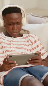Vertical video: Glancing left teen tapping gray tablet studying on couch, wearing headphones
