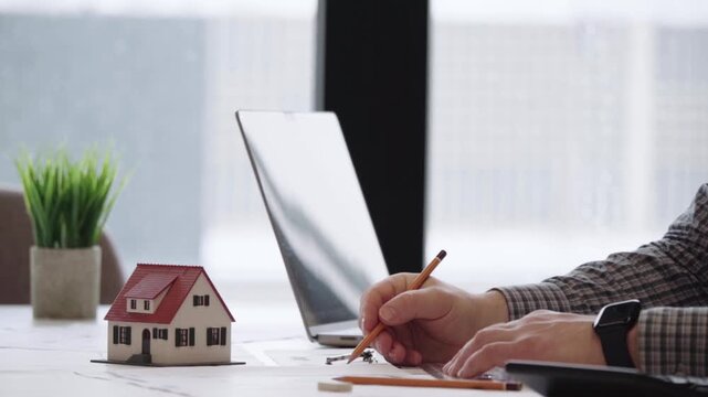 Hand writes beside laptop, small house model nearby. Focused, calm, precise. Office desk, natural light, minimalistic setting. Real estate planning, financial analysis, or startup concept