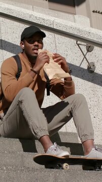 Vertical video: Opening bag, man in cap pulling food and eating on steps with skateboard for lunch