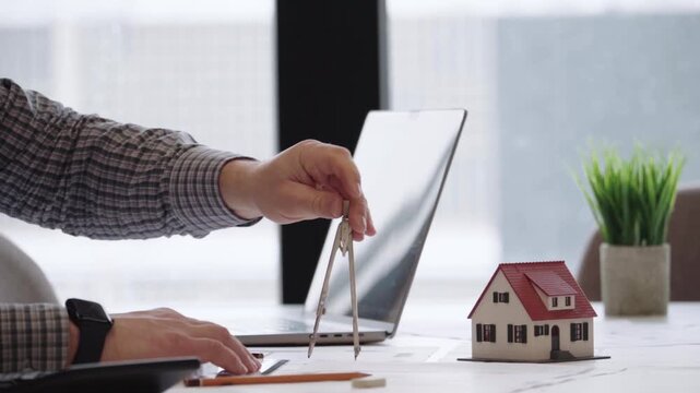 Hand holds compass near miniature house model. Laptop sits beside desk, laptop open. Person in plaid shirt draws plans for house. Work space has potted green plant
