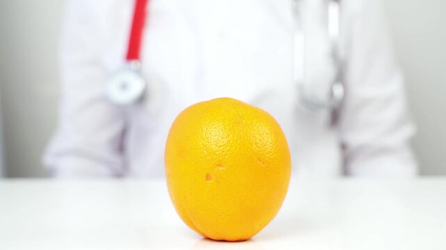 Doctor holds orange on white table. Orange sits center, vibrant, textured peel. Doctor's face hidden, focused on fruit. Healthcare professional suggests nutritional value. Ideal for health, wellness