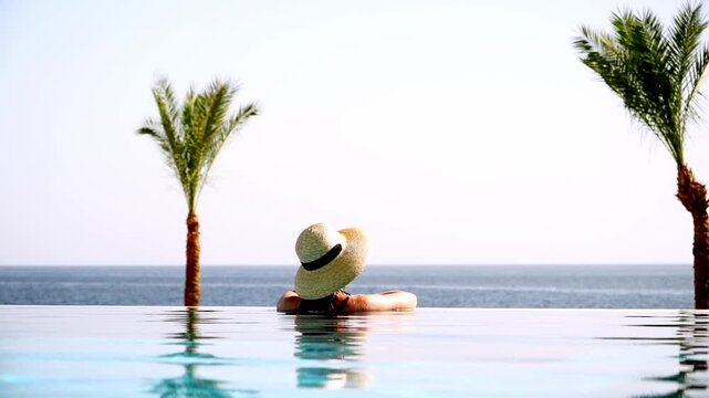 Woman relaxes in infinity pool, hat on head. Palm trees frame serene ocean view. Sunlight glints off clear water surface. Stillness evokes calm, peaceful escape. Ideal for travel, vacation