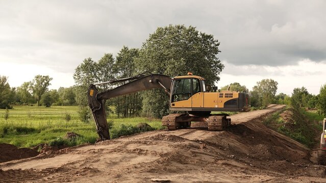 Zdjęcie przedstawia ż&oacute;łtą koparkę gąsienicową pracującą przy formowaniu ziemnego wału lub drogi. W tle widoczne są zielone drzewa, pola oraz zachmurzone niebo.