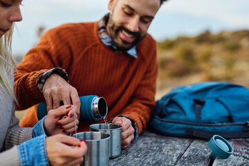 Fototapeta premium Hiking couple enjoys nature taking a break with water bottle or hot drink tea at the table in a lush forest during a sunny day