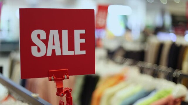 Close up of red sale sign board in department store of shopping mall. Bright clearance banner hanging over blurred clothes hangers on background. Seasonal discount offer and consumerism concept.