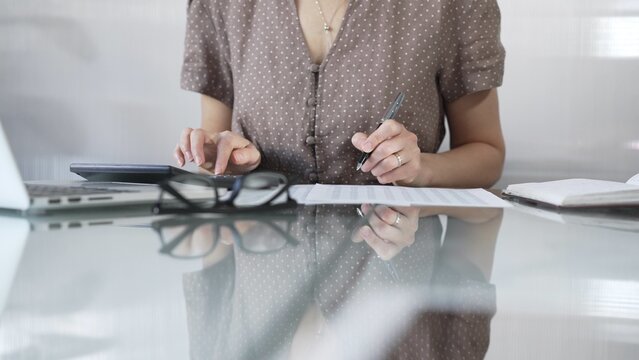 Businesswoman accountant, wearing brawn dotted blouse, is using a calculator to crunch numbers and jotting down meticulous notes, at sleek glass desk in a contemporary office. Taxes, audit in business