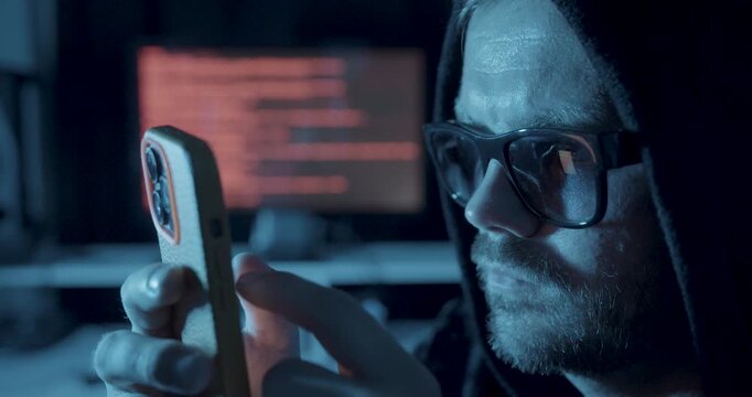 Hooded man using a smartphone in a dark room with computer code in the background, representing cybercrime, hacking, phishing, and digital security threats.