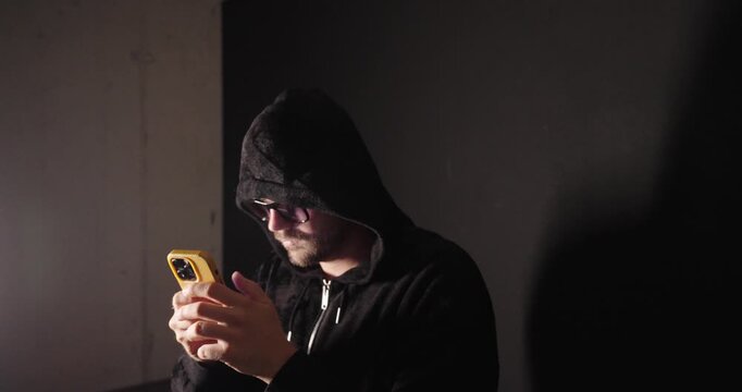 Hooded man using a smartphone in a dark room, representing cybercrime, phishing, hacking, online fraud, and digital security threats.