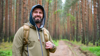 Man with backpack stands in pine forest and looks toward camera during hiking trip. Active recreation, exploration and travel lifestyle © Dmitrii Masquerade