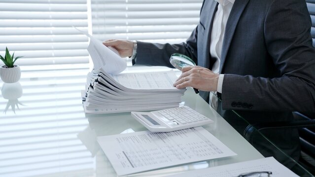Professional businessman using a magnifying glass to meticulously examine financial reports and paper documents on a desk, analyzing data for audit or investigation purposes