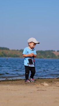 Lovely kid walks by the sandy river bank with no other people around. Toddler picks up a stone and throws it into water. Vertical video