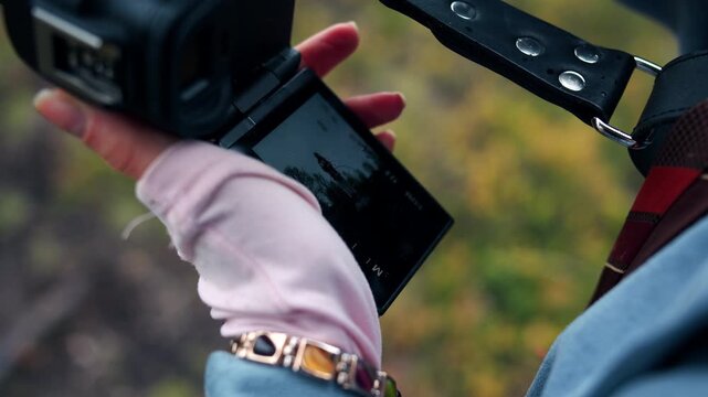 Close-up of a left female hand in a pale pink glove holding a mirrorless camera attached to a leather belt wit flip-out LCD screen tilted toward the viewer. The photographer is taking series of photos