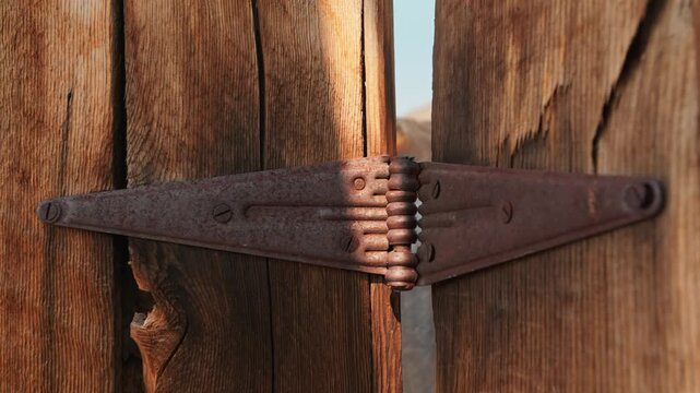 Old abandoned ghost town in America featuring a rusty iron hinge mounted on weathered wooden planks. Close-up reveals aged metal hardware with oxidation and textured grain patterns in warm sunlight.
