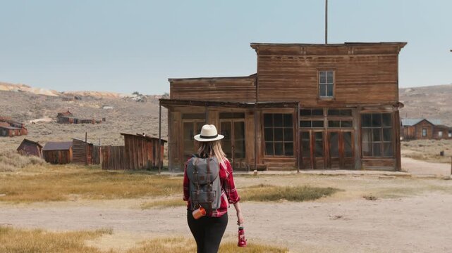 Young woman traveler with backpack and straw hat walks toward weathered wooden saloon building in abandoned Western ghost town, golden desert grass and rustic structures under clear blue sky.