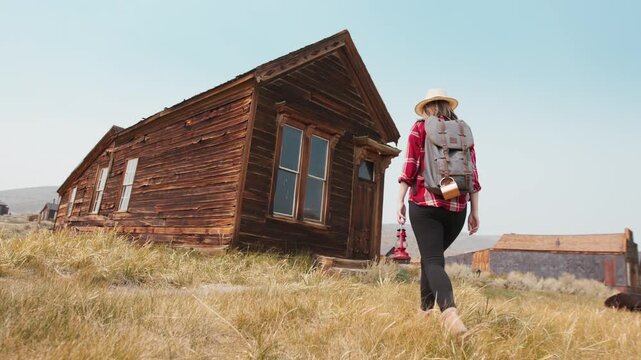 Girl walks through old abandoned ghost town in America exploring weathered wooden buildings. Woman with backpack and lantern discovers historic ruins in dry grassland under clear blue sky.