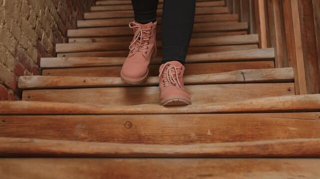 Close-up view of woman wearing pink lace-up leather boots and dark jeans descending rustic wooden log staircase in narrow corridor with weathered brick wall in old historic building interior.