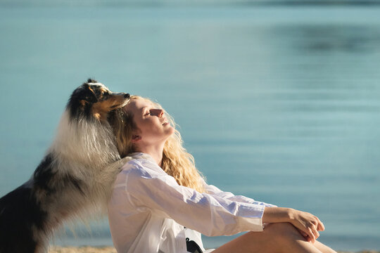A smiling woman cuddles her Shetland Sheepdog while relaxing beside a peaceful mountain lake. Warm lifestyle portrait expressing affection, trust, and emotional connection.
