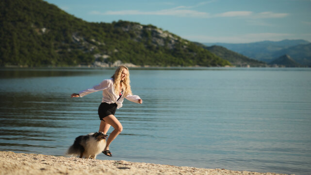 A woman exercises with her Shetland Sheepdog on a sandy lakeshore overlooking calm water and mountains. Inspiring outdoor scene about friendship, fitness, and travel.