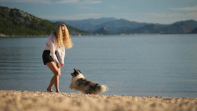 A woman enjoys training and playing with her Shetland Sheepdog beside a calm mountain lake. Natural lifestyle image expressing trust, companionship, and active outdoor recreation.