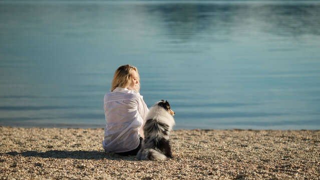 A woman and her Shetland Sheepdog sit together beside a calm lake surrounded by mountains. Peaceful outdoor lifestyle scene celebrating friendship and nature.