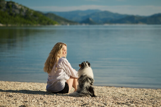 A woman sits beside her Shetland Sheepdog on a quiet lakeshore enjoying a peaceful moment together. Lifestyle image expressing friendship, trust, and outdoor relaxation.